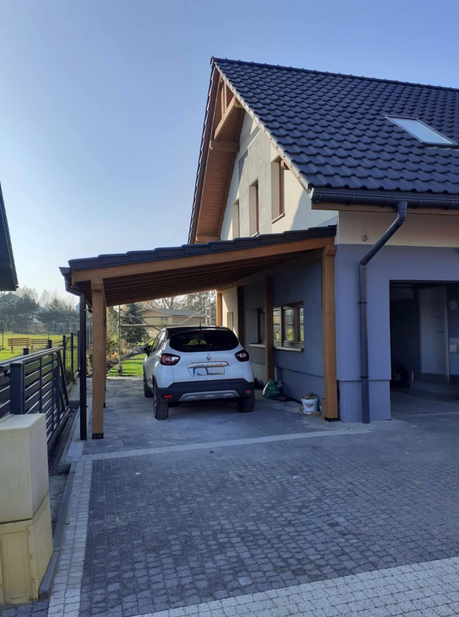 Nowoczesna wiata garażowa z drewna i ciemnej dachówki, przylegająca do domu z szarą elewacją. Biały samochód zaparkowany wewnątrz. Kostka brukowa na podjeździe.