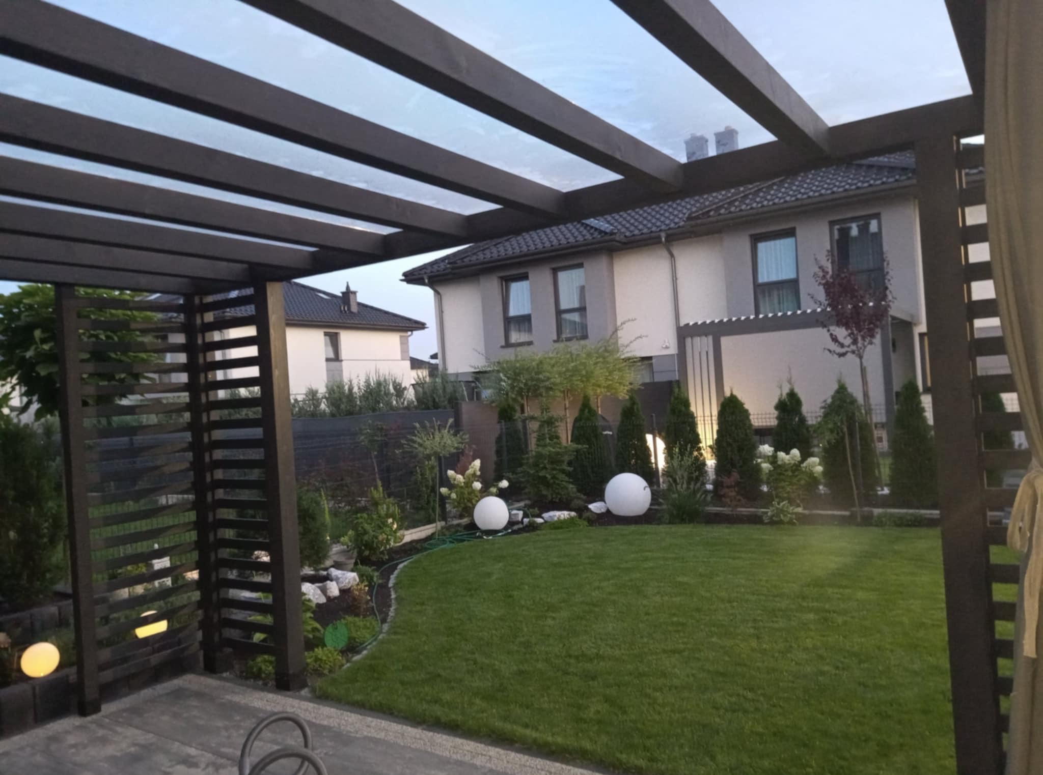 Nowoczesna pergola z ciemnego drewna z przezroczystym zadaszeniem, oświetlony ogród z kulistymi lampami, w tle dom jednorodzinny. Elegancki taras.