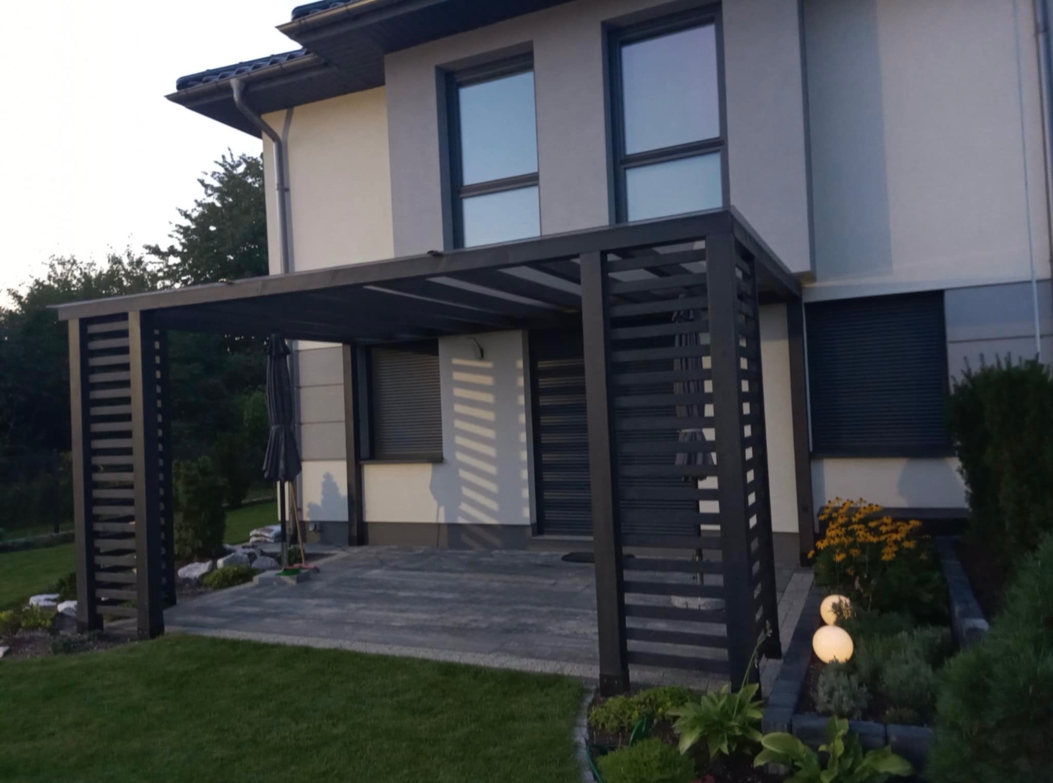 Nowoczesna, grafitowa pergola z ażurowymi ścianami, przylegająca do jasnego domu z szarymi oknami. Widoczne oświetlenie ogrodowe i zadbany trawnik.