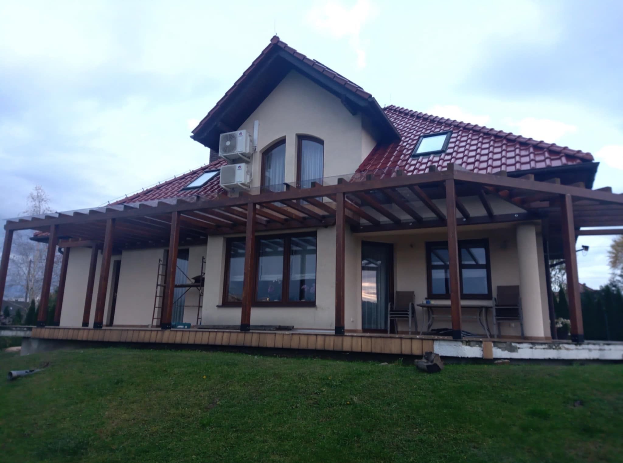 Dom z drewnianą pergolą tarasową w trakcie budowy, widoczne rusztowanie i niedokończone elementy wykończeniowe. Tło: pochmurne niebo i trawnik.