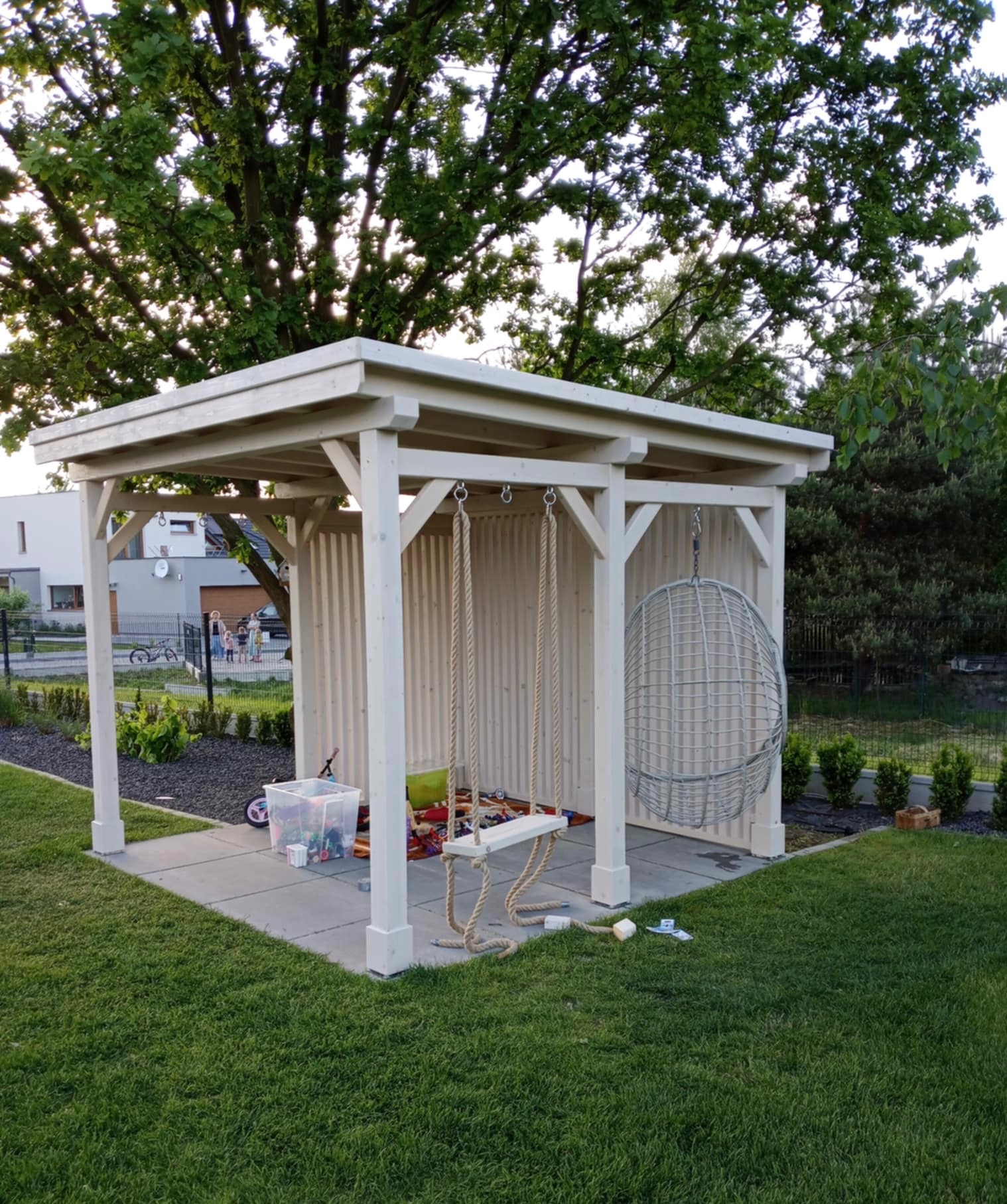 Biała, drewniana altana ogrodowa z huśtawką i wiszącym fotelem, otoczona zielonym trawnikiem i drzewami. W tle widoczny dom jednorodzinny.