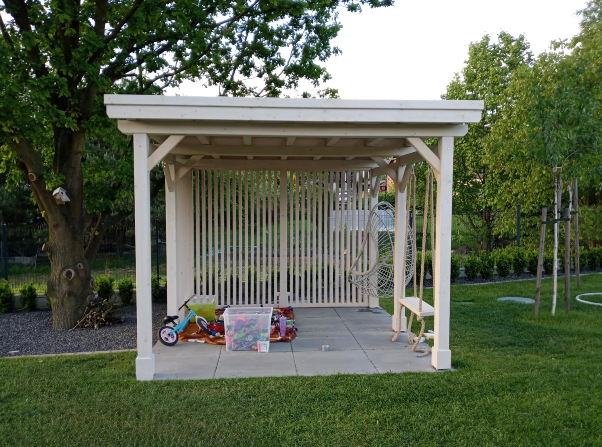 Biała, drewniana altana w ogrodzie z huśtawką, rowerkiem i zabawkami. Widok na zewnątrz przez ażurowe ściany. Trawa, drzewa i krzewy w tle.