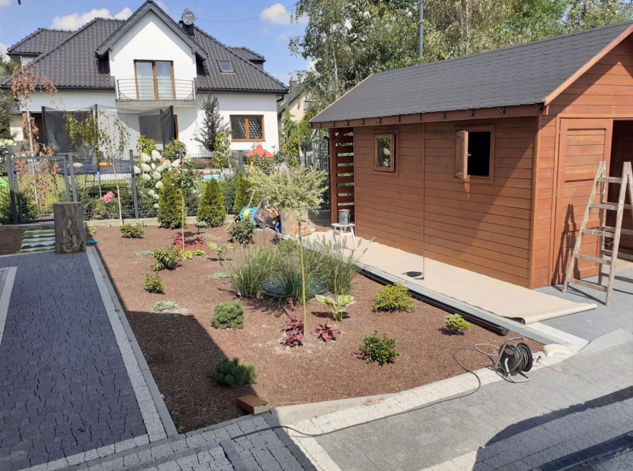 Drewniana altana z otwartymi oknami i drzwiami, usytuowana w ogrodzie z nowo nasadzonymi roślinami i wybrukowanym podjazdem. W tle widoczny dom.