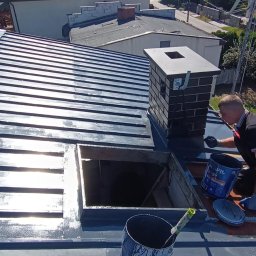 Oleksii Levytskyi - Malowanie metalowego dachu na szaro, widoczny komin z cegły, puszka z farbą i pracownik w rękawicach. Ujęcie z góry, w tle zabudowa jednorodzinna.