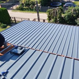 Oleksii Levytskyi - Malowanie dachu z blachy trapezowej na szaro. Widoczny wałek i kuweta z farbą. W tle domy i zieleń. Ujęcie z góry, ukazujące strukturę blachy i etap prac malarskich.