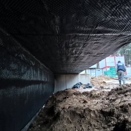 Oleksii Levytskyi - Ochrona fundamentów budynku folią kubełkową, widoczny grunt i fragment konstrukcji. Prace ziemne w toku, w tle kontenery budowlane i osoba w odzieży roboczej.