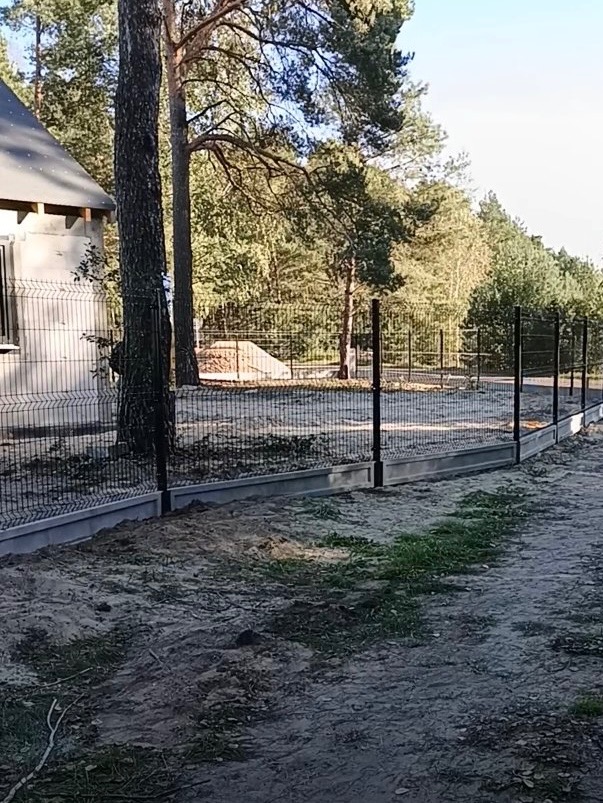 Nowoczesne ogrodzenie panelowe z podmurówką betonową, czarne słupki, widok na las w tle, dom w budowie po lewej stronie kadru, słoneczny dzień.