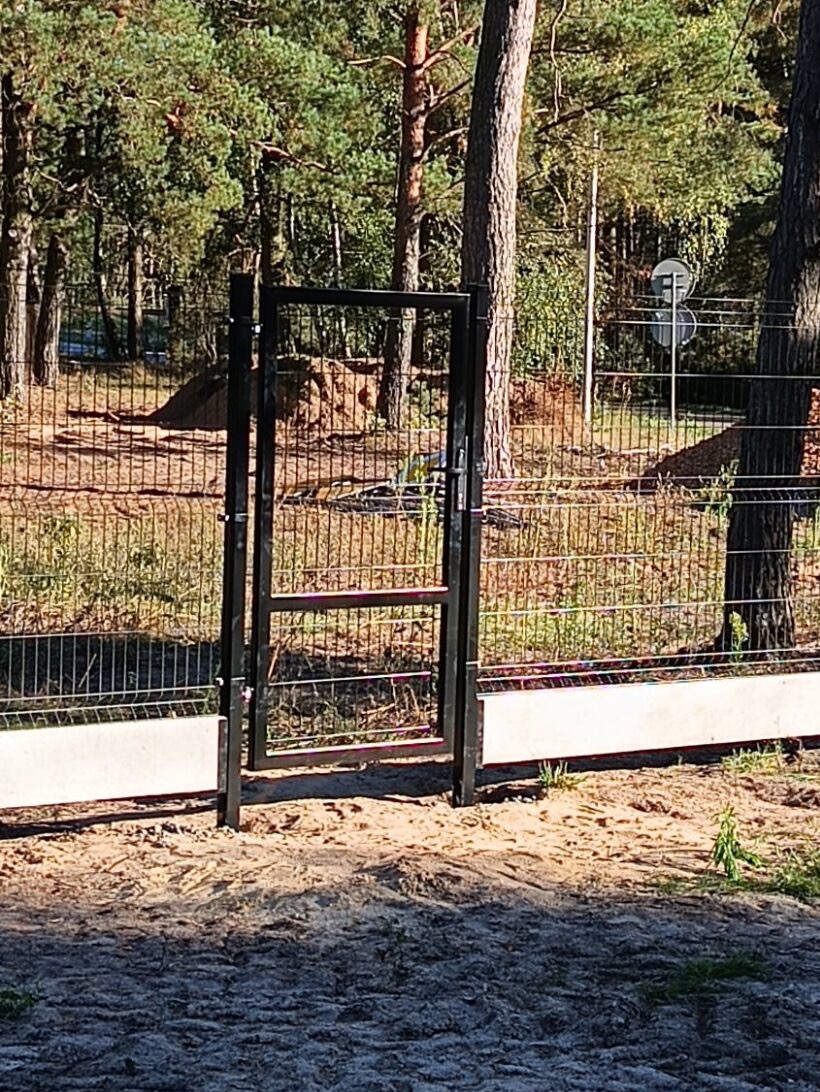 Nowoczesna furtka z metalową ramą i siatką, osadzona na betonowej podmurówce. W tle las i znak drogowy. Ujęcie w słoneczny dzień.