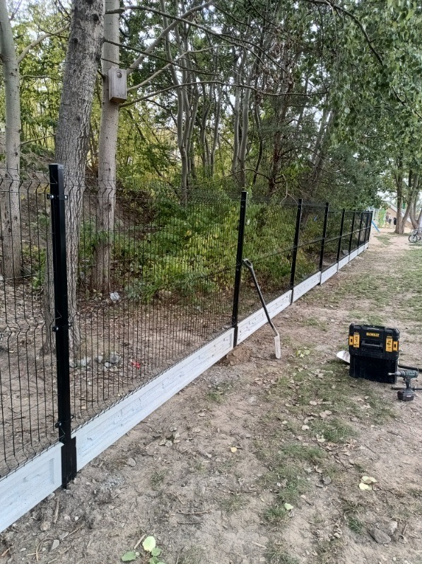 Częściowo ukończone ogrodzenie panelowe z czarnymi słupkami i betonową podmurówką, narzędzia pracy obok, widok na zalesiony teren w tle.
