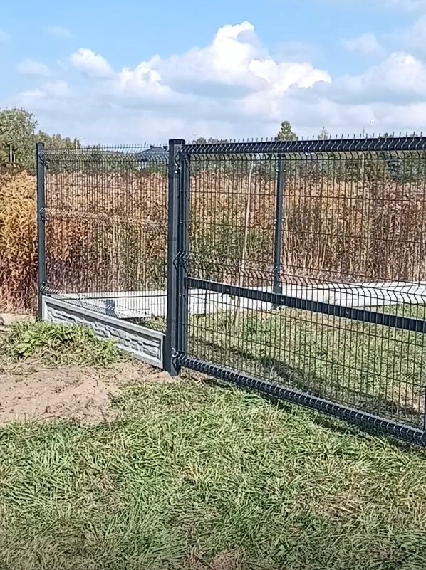 Nowoczesne ogrodzenie panelowe w kolorze grafitowym, z podmurówką imitującą kamień, na tle zielonej trawy i wysokich traw. Widok z bliska, słoneczny dzień.