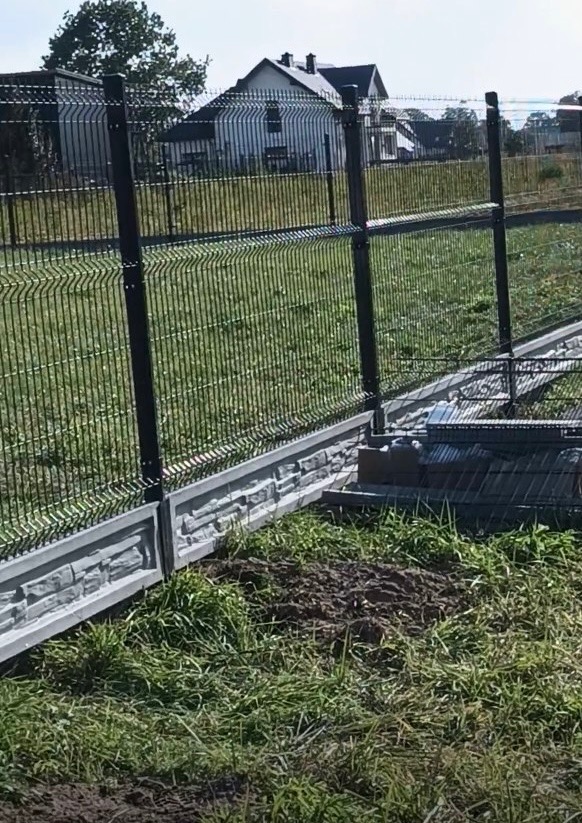 Panelowe ogrodzenie z podmurówką imitującą kamień, na tle zielonej trawy i domu. Widoczne słupki i siatka ogrodzeniowa. Ujęcie z bliska, podkreślające detale.