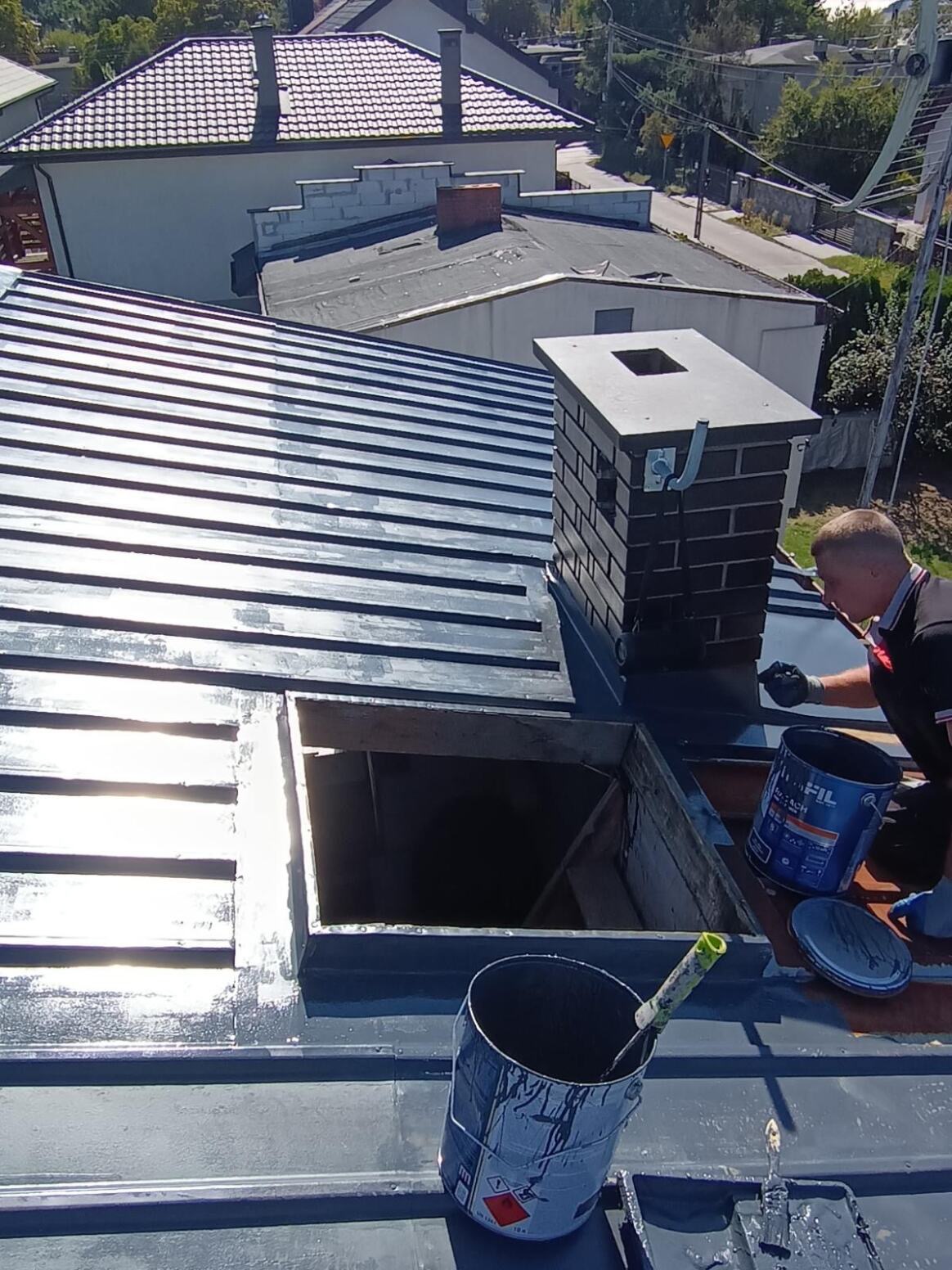 Malowanie metalowego dachu na szaro, widoczny komin z cegły, puszka z farbą i pracownik w rękawicach. Ujęcie z góry, w tle zabudowa jednorodzinna.