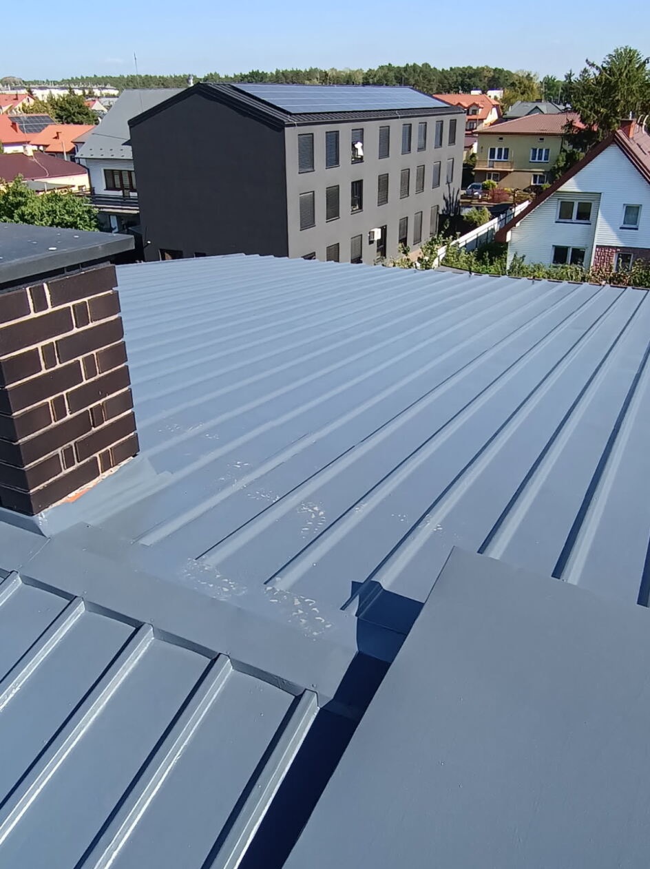 Szary, blaszany dach z kominem z cegły na tle zabudowy miejskiej w słoneczny dzień. Widoczne panele słoneczne na budynku w tle.