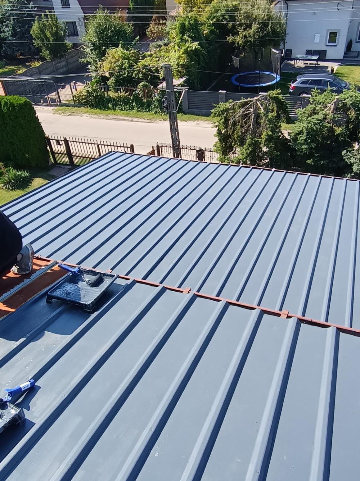 Malowanie dachu z blachy trapezowej na szaro. Widoczny wałek i kuweta z farbą. W tle domy i zieleń. Ujęcie z góry, ukazujące strukturę blachy i etap prac malarskich.