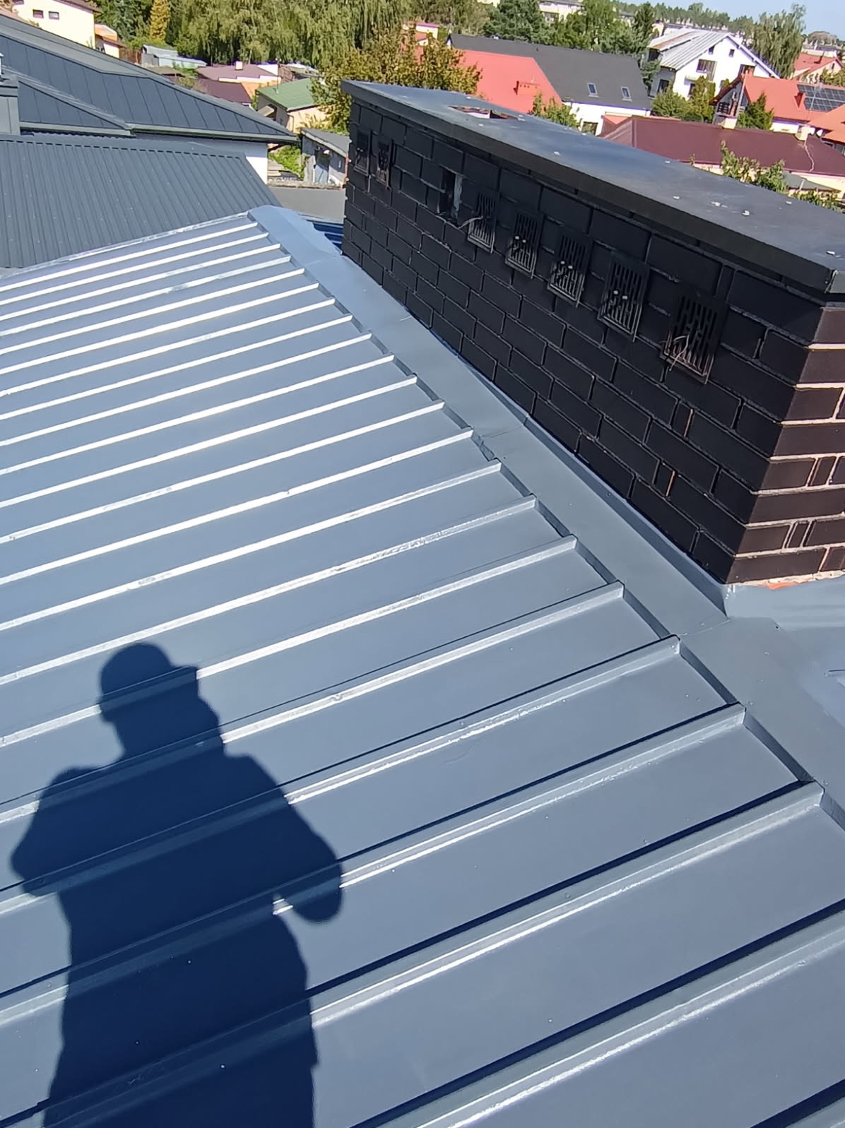 Szary blaszany dach z widocznym kominem z cegły i cieniem osoby, ukazujący detale wykończenia i strukturę blachy na tle zabudowań.