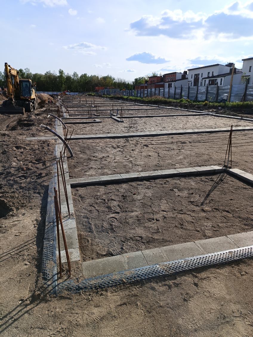 Przygotowanie terenu pod fundamenty ogrodzenia z użyciem szalunków i zbrojenia. W tle widoczny sprzęt budowlany i zabudowa mieszkalna.