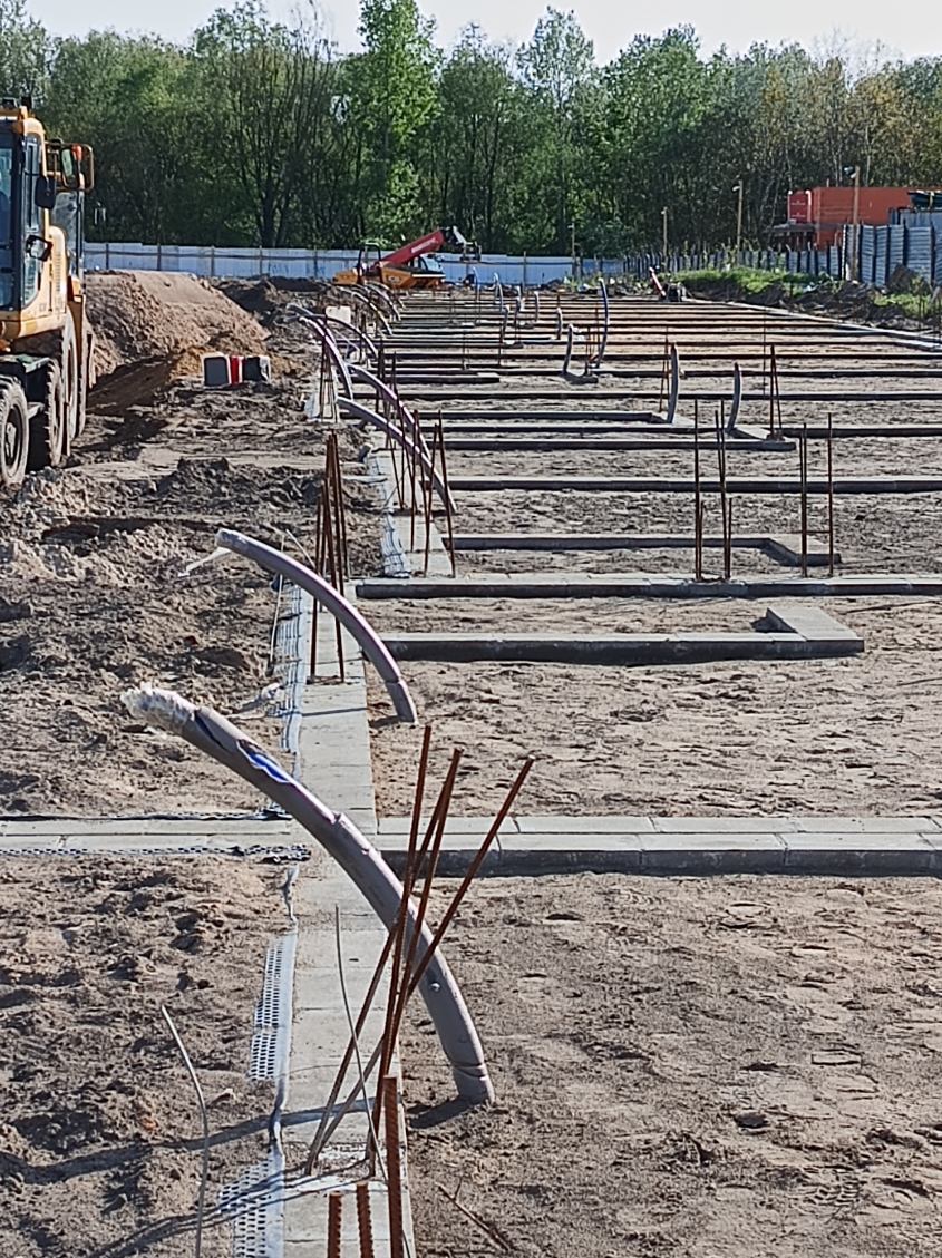 Fundamenty pod budowę z widocznymi zbrojeniami, rurami i elementami szalunków. W tle sprzęt budowlany i drzewa.