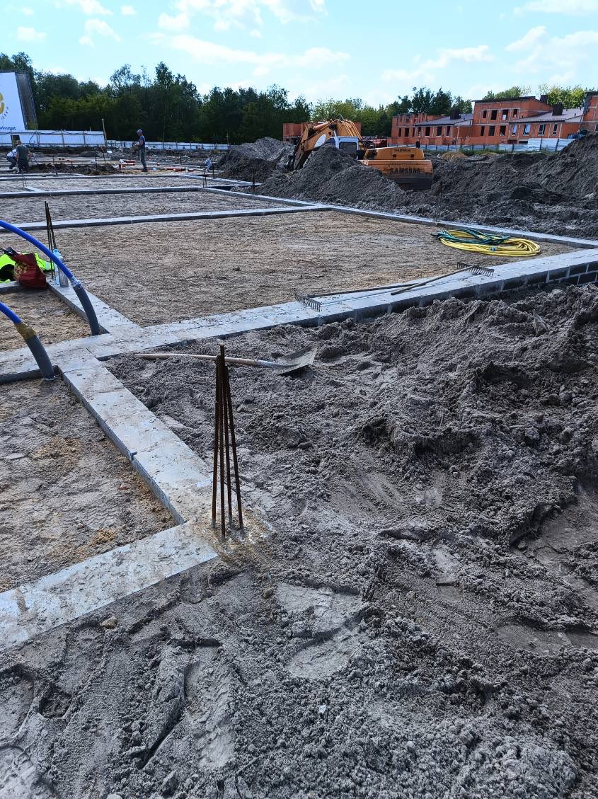 Fundamenty domu w trakcie budowy, z widocznymi zbrojeniami, betonowymi bloczkami i sprzętem budowlanym w tle, w słoneczny dzień.