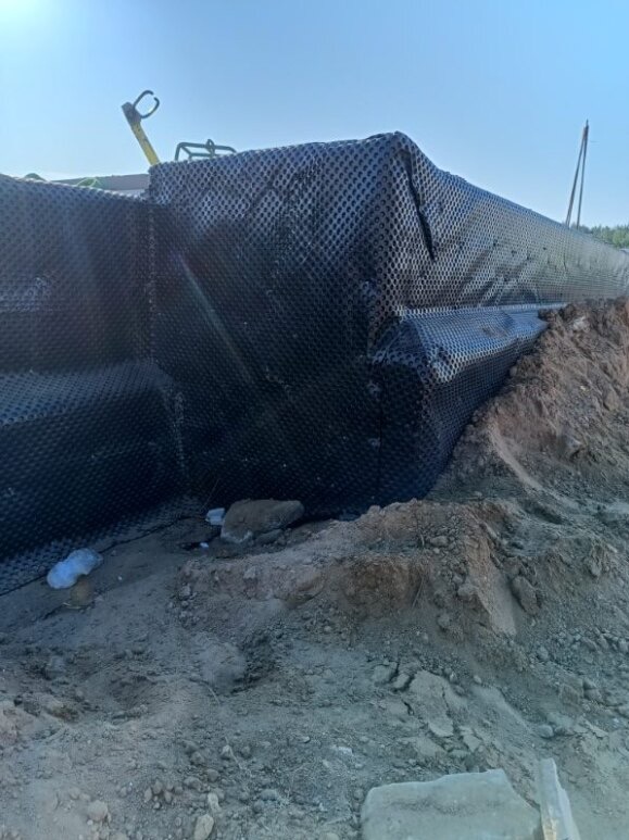 Czarna folia kubełkowa zabezpieczająca fundament budynku przed wilgocią, widoczna z bliska na tle ziemi i nieba. Etap prac budowlanych.