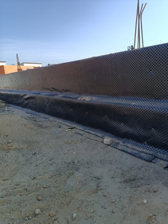 Ochrona fundamentów budynku w trakcie budowy: czarna folia kubełkowa zabezpiecza ścianę przed wilgocią. W tle widoczne zbrojenie i fragmenty konstrukcji.