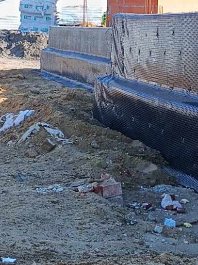 Fundament budynku z izolacją pionową w postaci folii kubełkowej, częściowo zasypany ziemią. Widoczne bloczki fundamentowe i worki z zaprawą w tle.