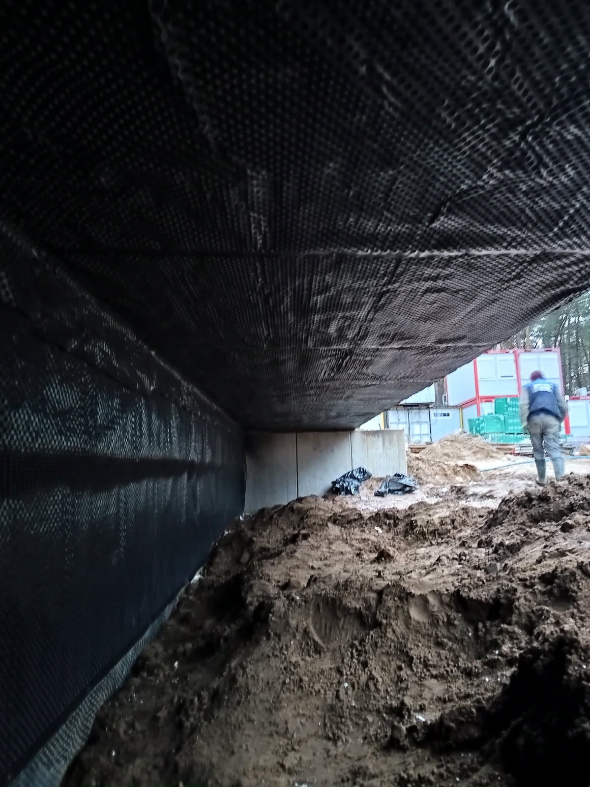 Ochrona fundamentów budynku folią kubełkową, widoczny grunt i fragment konstrukcji. Prace ziemne w toku, w tle kontenery budowlane i osoba w odzieży roboczej.