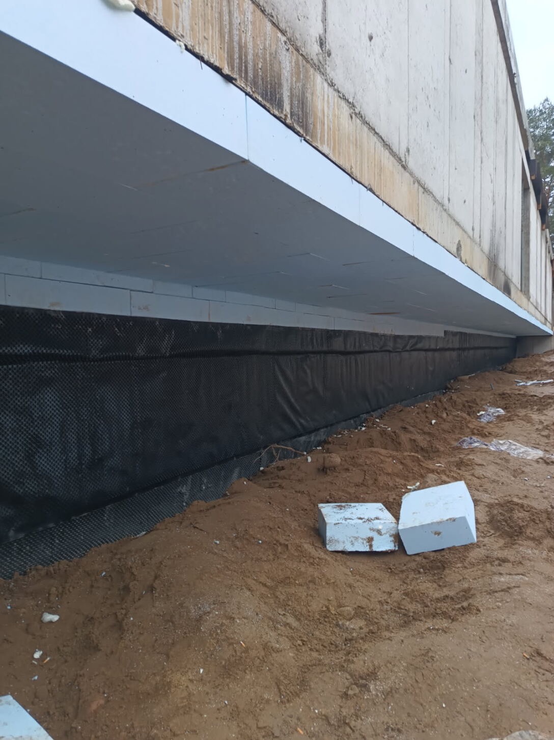 Ocieplenie fundamentów budynku w Warszawie, zabezpieczone folią kubełkową. Widoczne bloki styropianowe na ziemi.