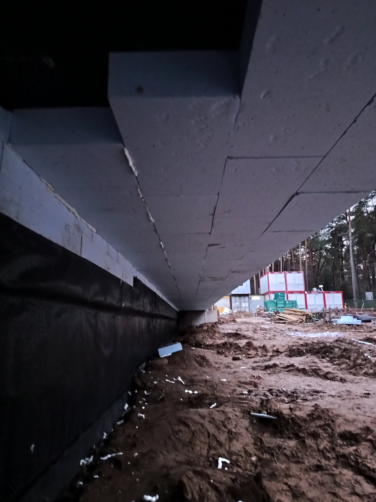 Izolacja fundamentów budynku w trakcie budowy, widok z dołu. Czarna folia izolacyjna na ścianie fundamentowej, styropianowe płyty izolacyjne. W tle kontenery budowlane i drzewa.