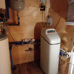 ECO-INSTAL - Biały zmiękczacz wody Viessmann w drewnianym pomieszczeniu technicznym, widoczne rury, zawory, zbiornik wyrównawczy i instalacja elektryczna.