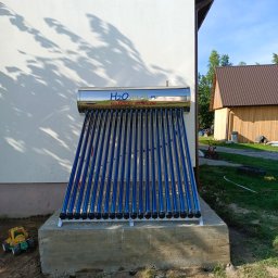 ECO-INSTAL - Zamontowany kolektor słoneczny H2O Solar na betonowym fundamencie przy białej ścianie budynku, z zabawką obok. Widok na budynek gospodarczy w tle.