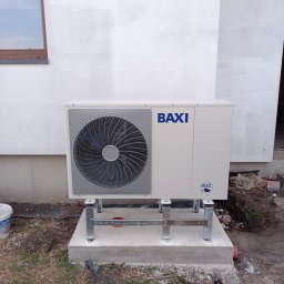 ECO-INSTAL - Biała pompa ciepła BAXI zamontowana na betonowym fundamencie przy ścianie budynku. Widoczne elementy montażowe i fragment otoczenia.