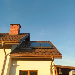 ECO-INSTAL - Dom z brązowym dachem i panelami solarnymi w słoneczny dzień. Widoczny komin z cegły, okno i rynna. Na dachu dwa panele solarne.