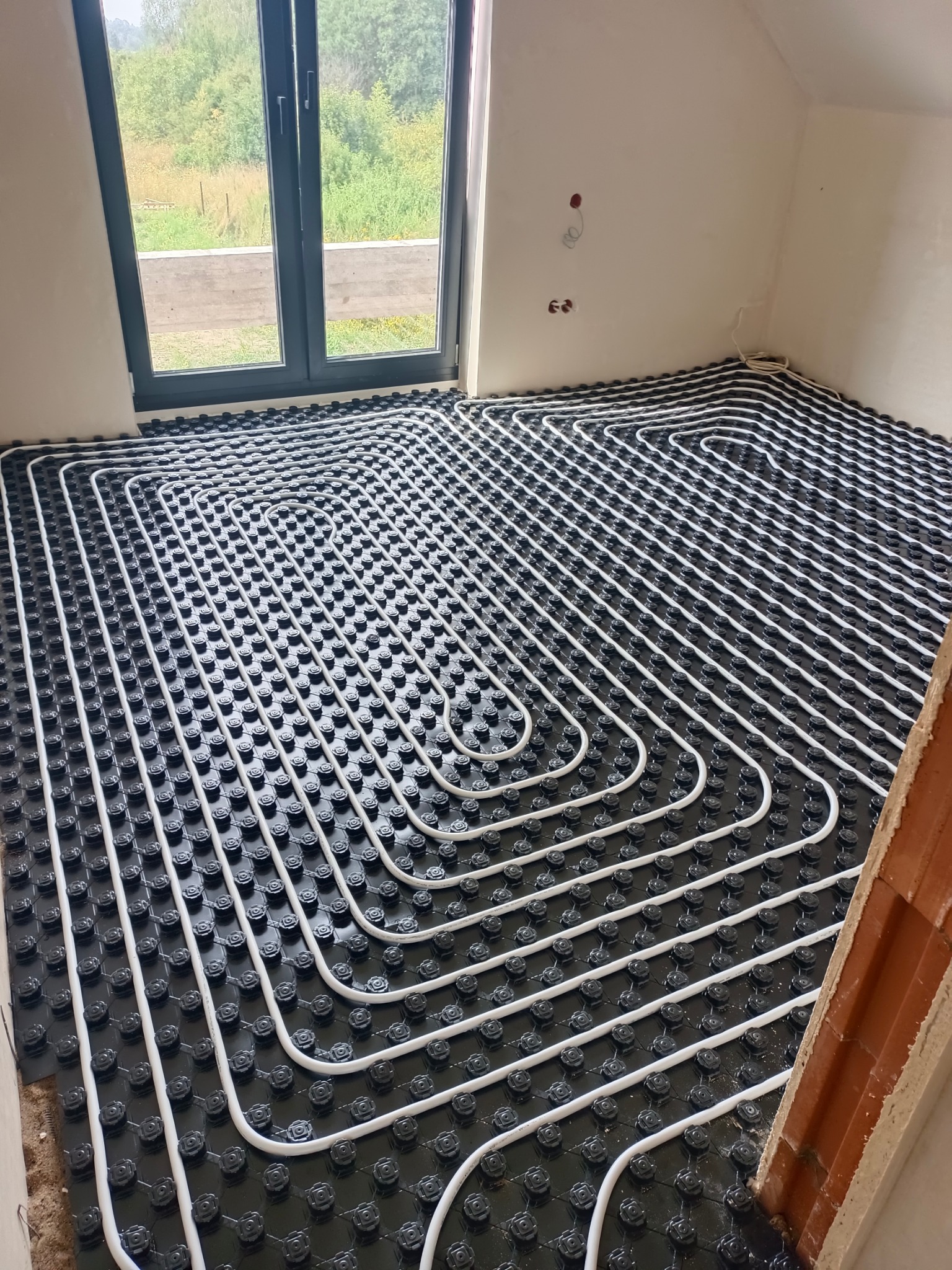 Białe rury ogrzewania podłogowego ułożone na czarnej macie w jasnym pomieszczeniu z oknem. Widoczny fragment ściany z cegły.