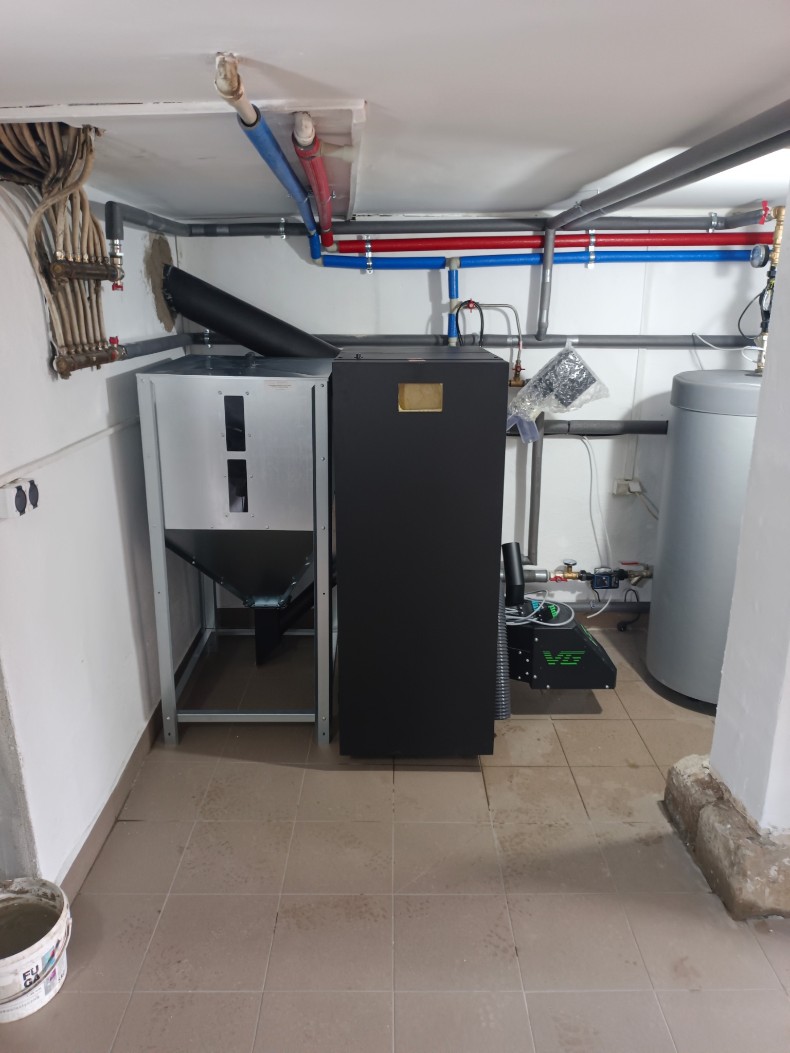 Nowoczesny piec na pellet z podajnikiem i zasobnikiem, zainstalowany w kotłowni. Widoczne rury instalacji grzewczej i zbiornik buforowy. Całość w jasnym pomieszczeniu.