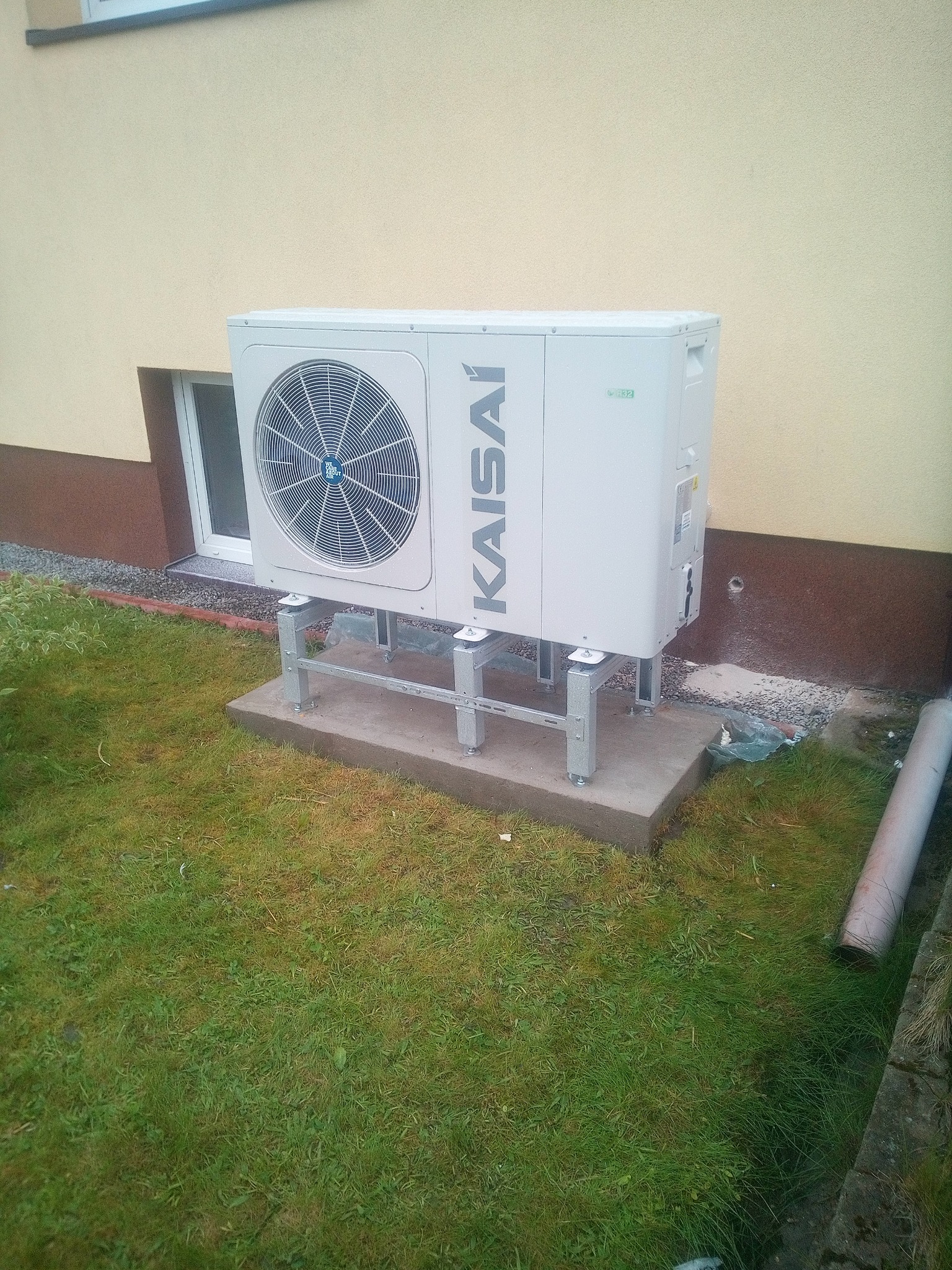 Biała pompa ciepła Kaisai zamontowana na betonowym fundamencie i metalowej konstrukcji, na tle trawnika i ściany budynku. Widoczny wentylator i logo.