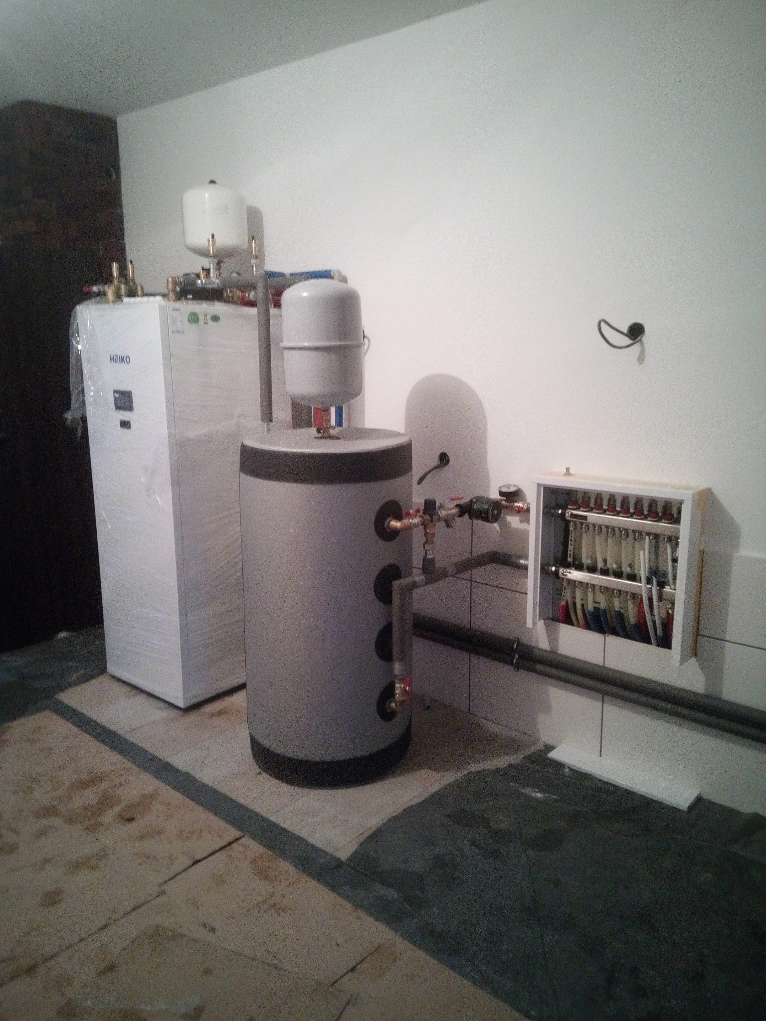 Nowoczesna instalacja grzewcza z zasobnikiem i rozdzielaczem w Tuczępy. Biały piec owinięty folią, szary zasobnik ciepłej wody, rozdzielacz w obudowie na ścianie.