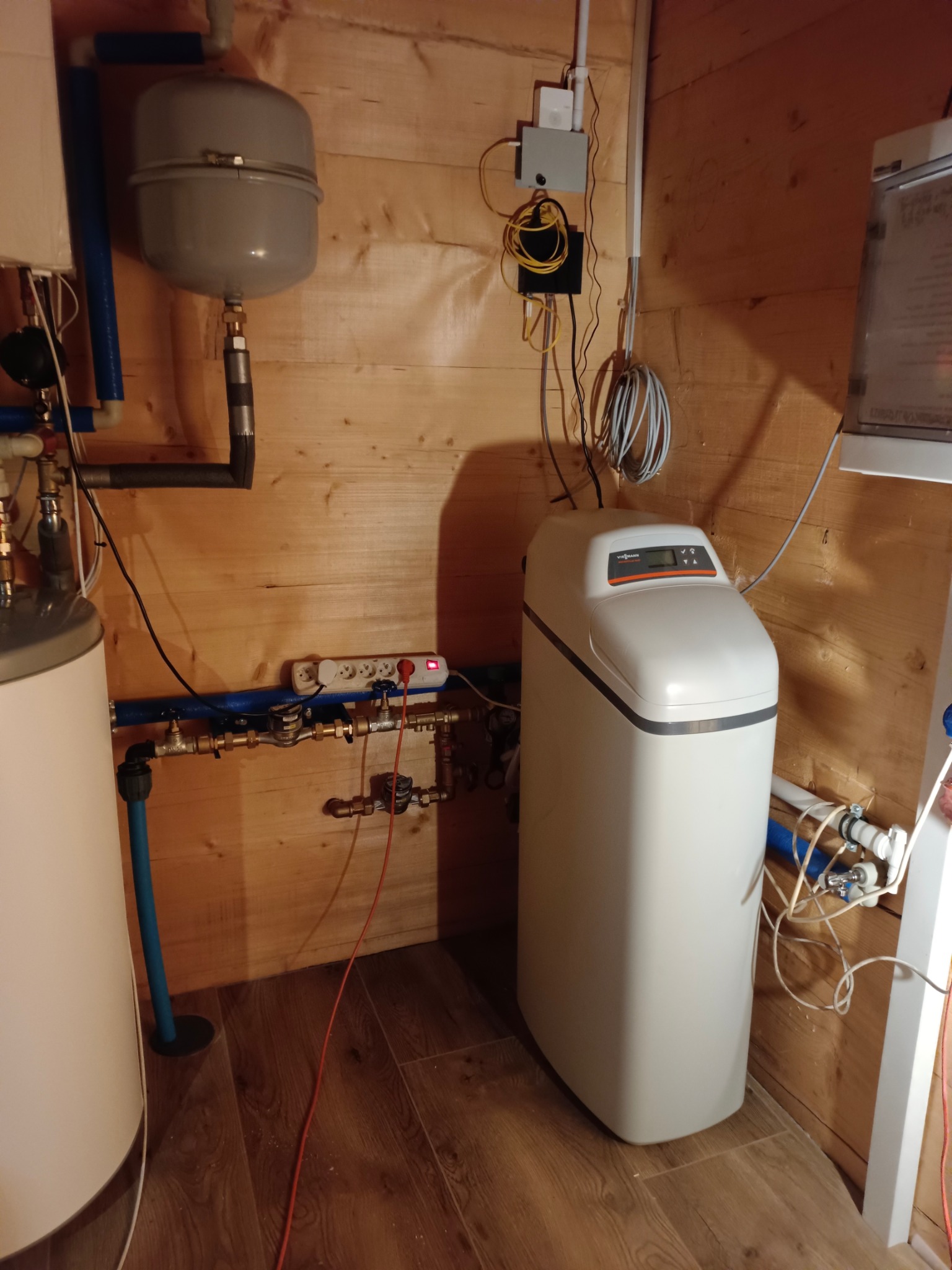 Biały zmiękczacz wody Viessmann w drewnianym pomieszczeniu technicznym, widoczne rury, zawory, zbiornik wyrównawczy i instalacja elektryczna.