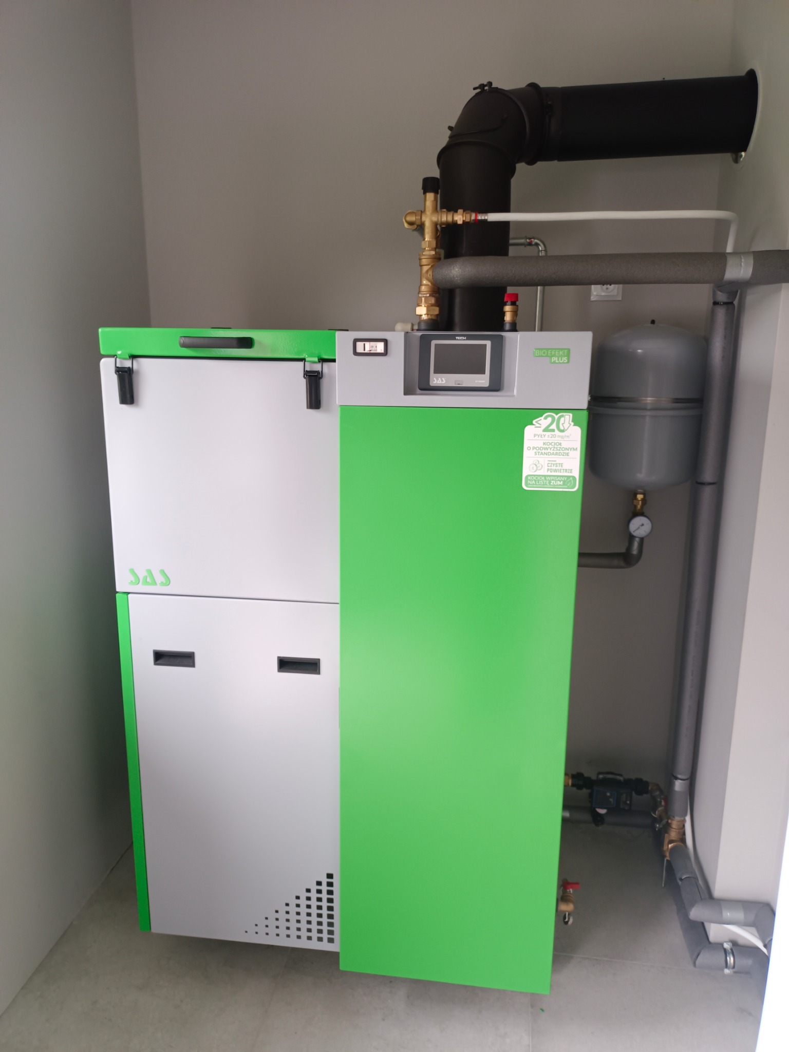 Nowoczesny piec SAS Bio EFEKT PLUS w kotłowni, z czarnym kominem i szarym zbiornikiem wyrównawczym. Widoczne rury i armatura. Ściany w kolorze szarym.
