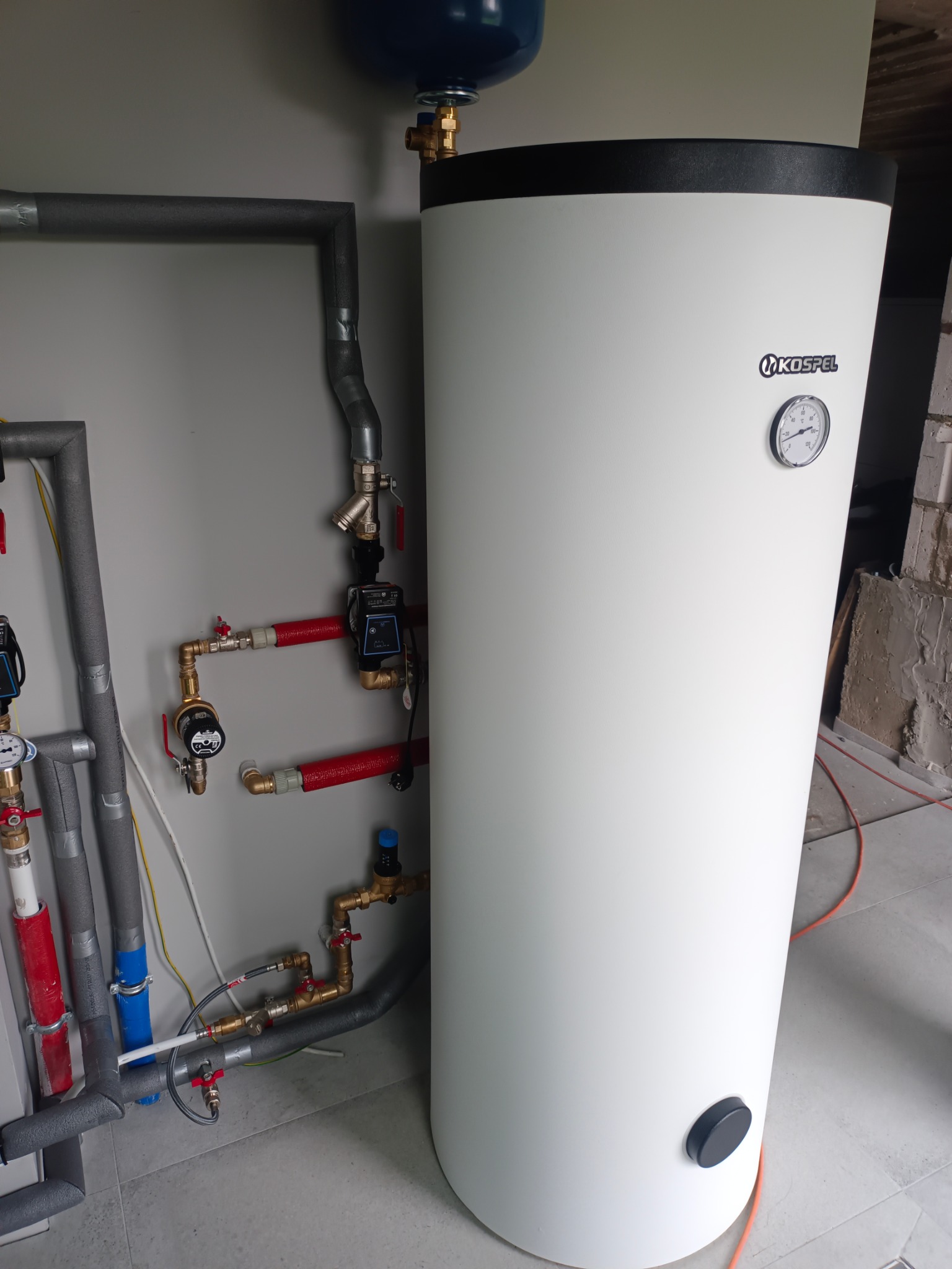 Biały, cylindryczny bojler Kospel z podłączonymi rurami instalacji grzewczej, widoczne zawory i pompy obiegowe. Szare ściany i podłoga z płytek.