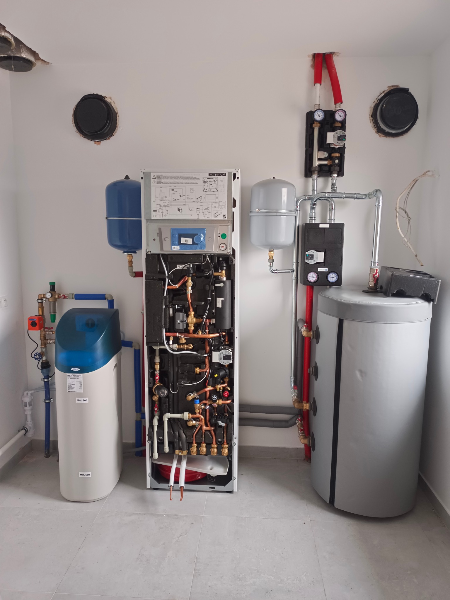 Kompletna instalacja hydrauliczna z piecem, zbiornikiem i zmiękczaczem wody. Widoczne rury miedziane, zawory i manometry. Białe ściany i jasna podłoga.