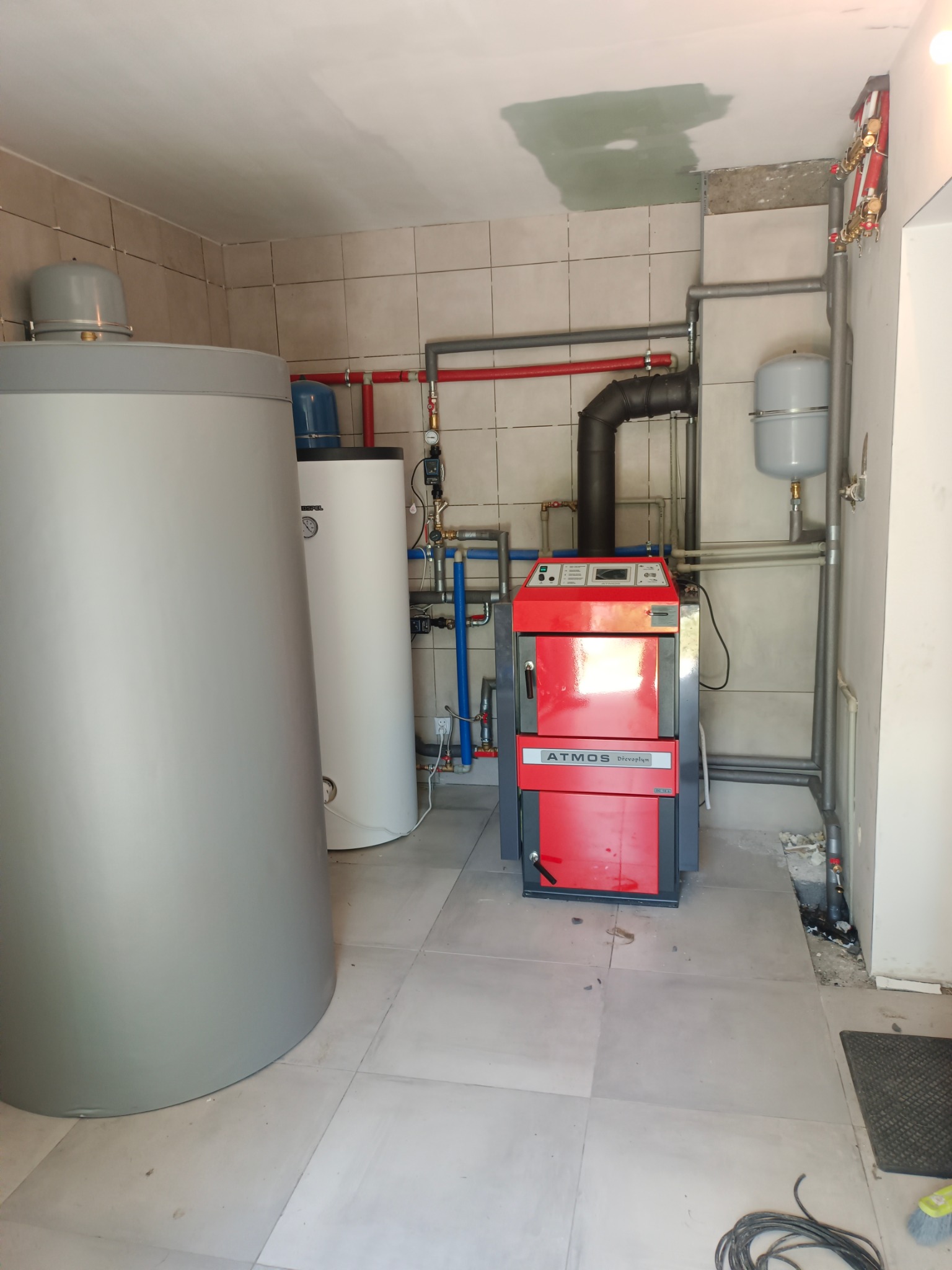 Zakończona instalacja kotła Atmos DrevoPlyn z zbiornikami buforowymi w kotłowni wyłożonej płytkami. Widoczne rury instalacji grzewczej i elementy armatury.