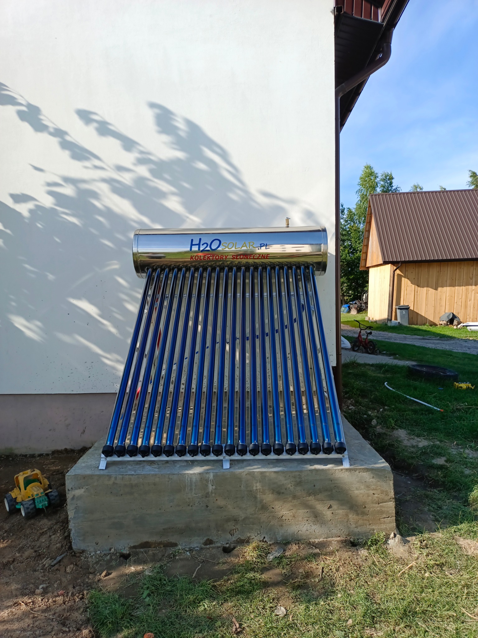 Zamontowany kolektor słoneczny H2O Solar na betonowym fundamencie przy białej ścianie budynku, z zabawką obok. Widok na budynek gospodarczy w tle.
