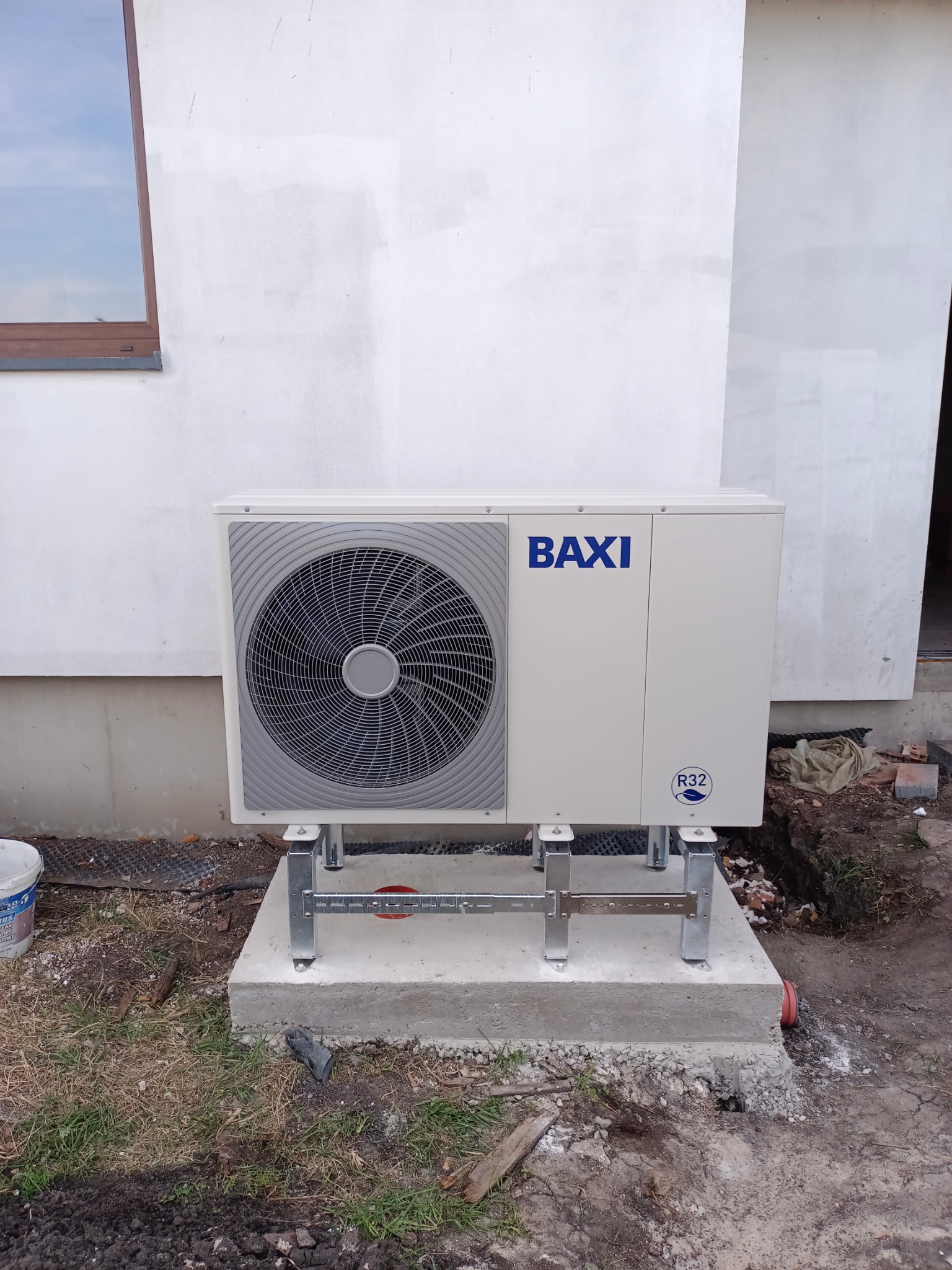Biała pompa ciepła BAXI zamontowana na betonowym fundamencie przy ścianie budynku. Widoczne elementy montażowe i fragment otoczenia.