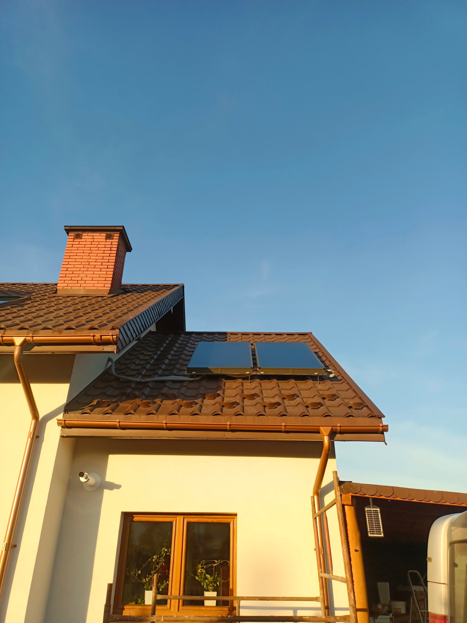 Dom z brązowym dachem i panelami solarnymi w słoneczny dzień. Widoczny komin z cegły, okno i rynna. Na dachu dwa panele solarne.