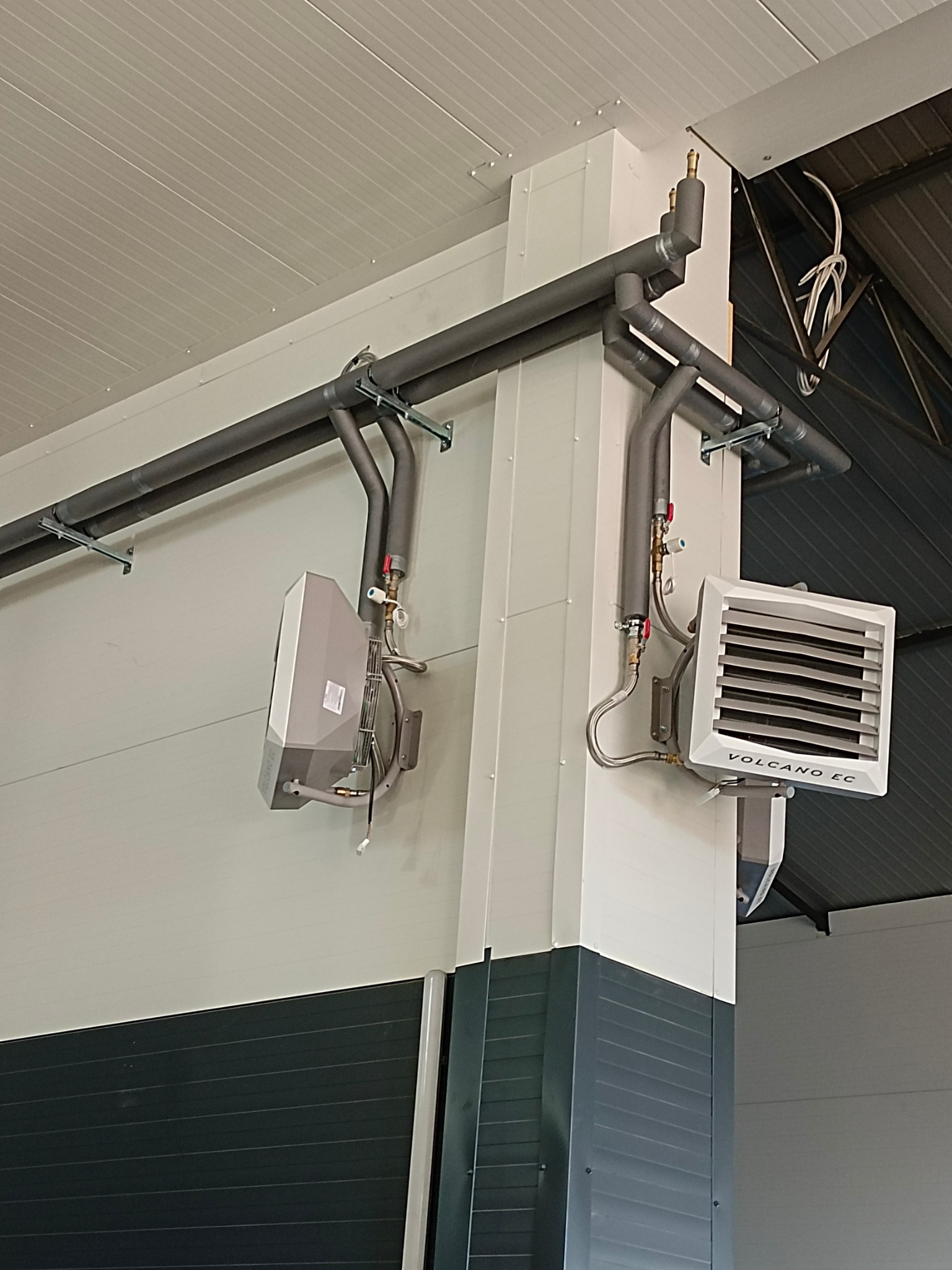 Dwa grzejniki Volcano EC zamontowane na ścianie, z widocznymi rurami doprowadzającymi wodę. Instalacja w hali przemysłowej, widoczne elementy konstrukcyjne budynku.