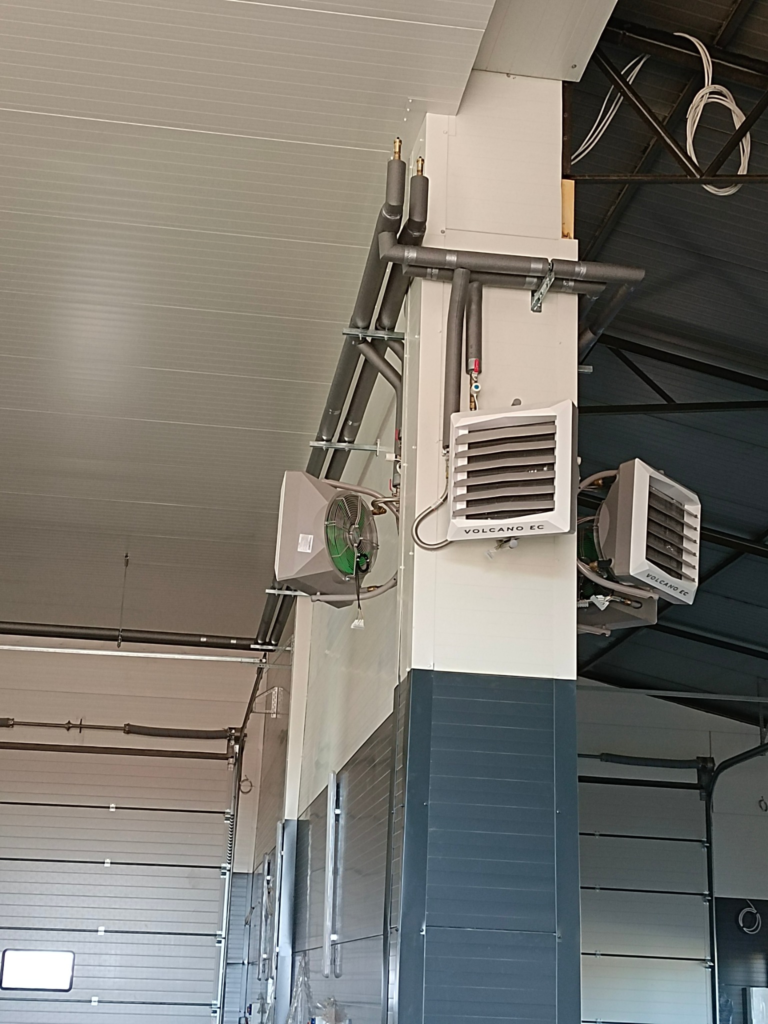 Zamontowane nagrzewnice VOLCANO EC z szarymi rurami w hali przemysłowej. Widoczne instalacje pod sufitem i fragment bramy garażowej. Ujęcie z dołu, podkreślające wysokość pomieszczenia.