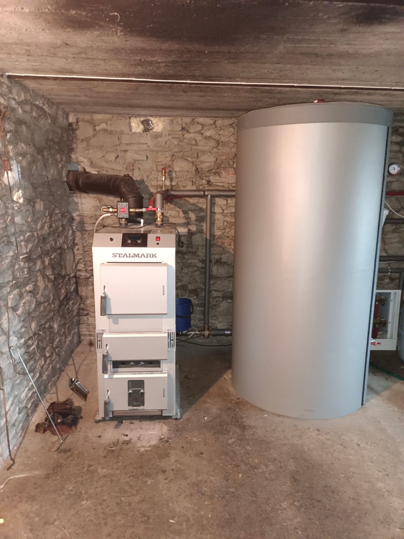 Nowoczesny piec Stalmark z zasobnikiem w kotłowni o kamiennych ścianach. Widoczne rury, armatura i akcesoria. Oświetlenie sufitowe.