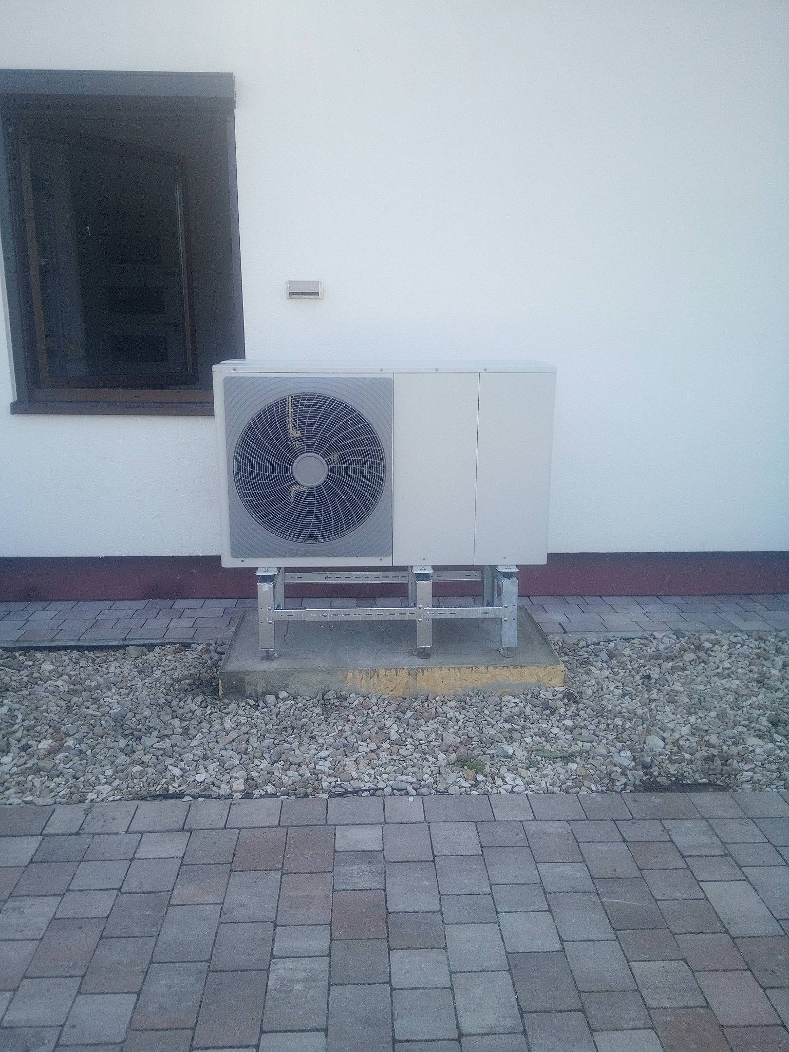 Biała pompa ciepła powietrze-woda na metalowej konstrukcji, zainstalowana przy ścianie budynku z oknem. Otoczenie wyłożone kostką i kamieniami.