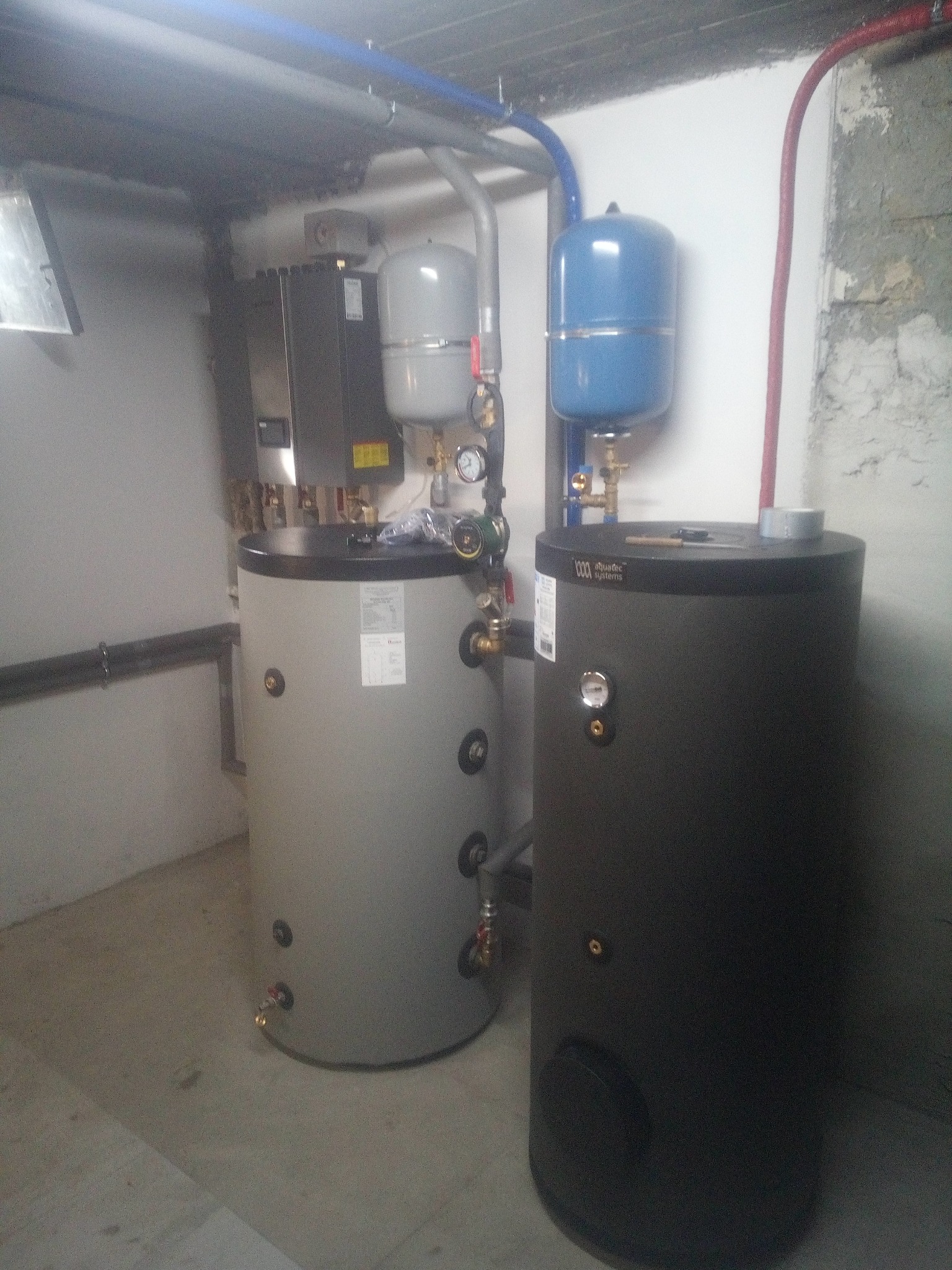 Dwa cylindryczne zbiorniki, szary i czarny, z podłączonymi rurami w kotłowni. Widoczne manometry i elementy instalacji.