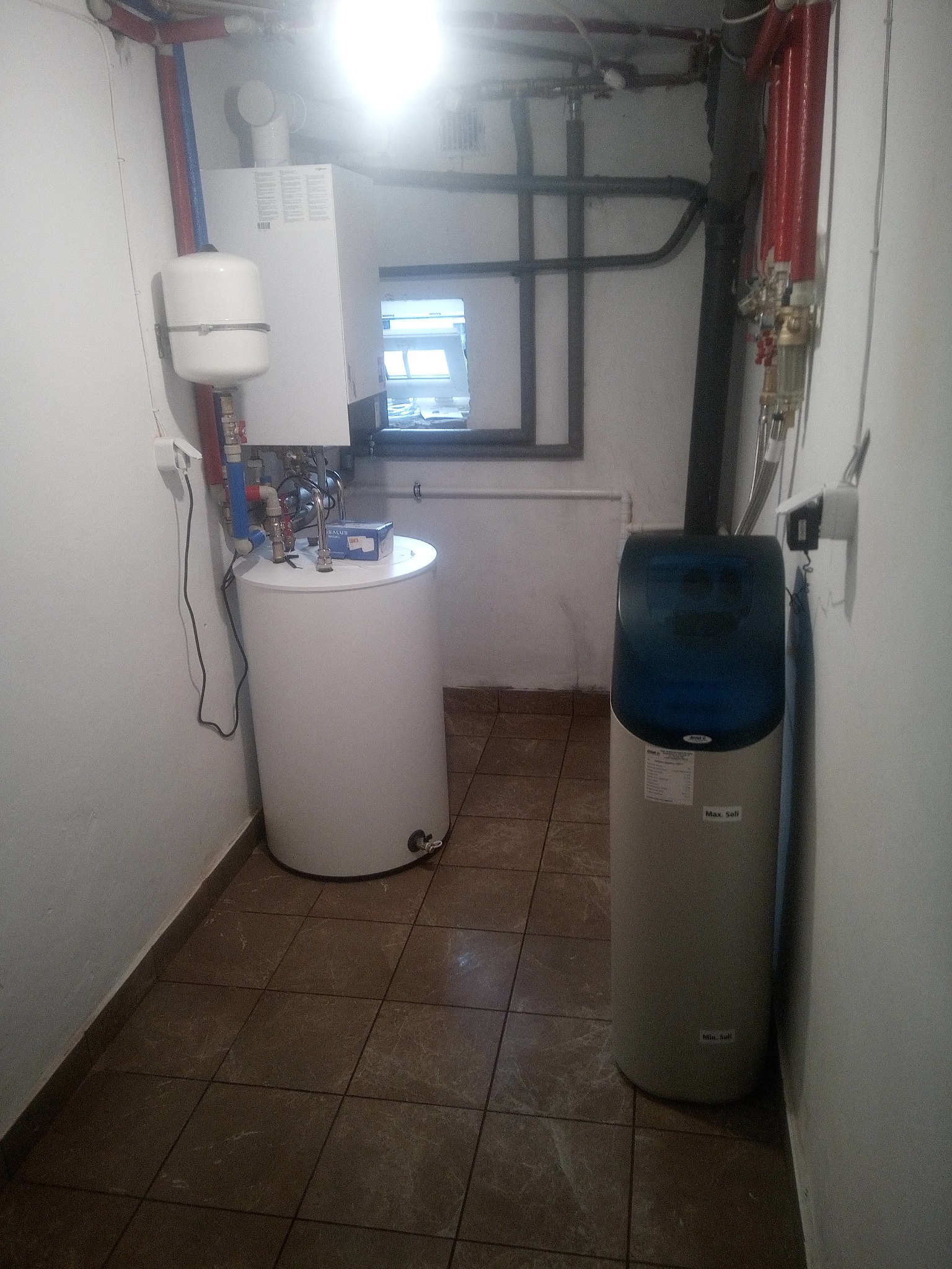 Kotłownia z piecem gazowym, zasobnikiem CWU i zmiękczaczem wody. Widoczne rury instalacji CO i CWU. Brązowe płytki na podłodze. Okno w tle.